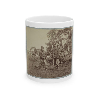 Sepia-toned print of two soldiers with bayoneted rifles standing in a field before wagon wheels and a fallen log.