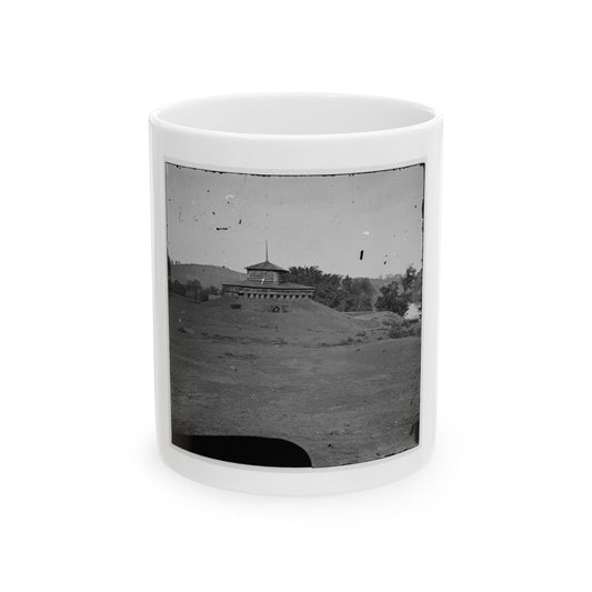 Black-and-white view of a small log blockhouse with a pyramidal roof on a grassy mound, with hills behind.