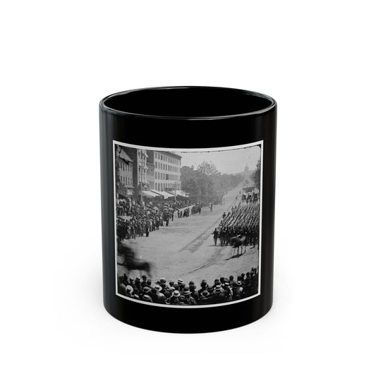 Black and white 19th-century view of marching soldiers with upright rifles on a broad avenue lined with spectators.