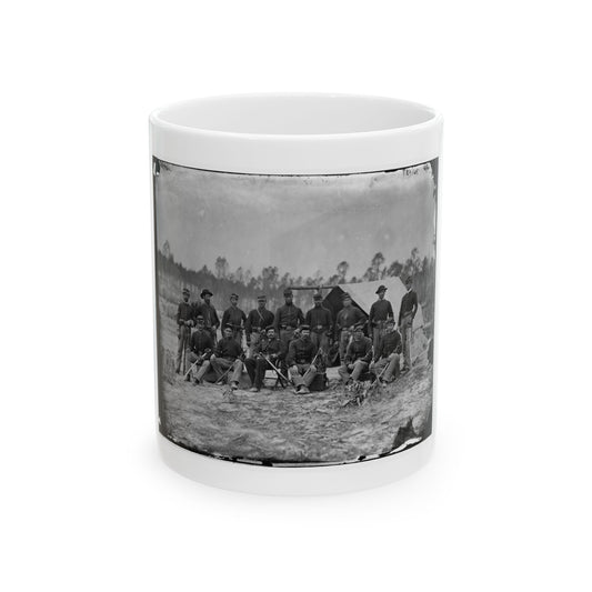 Black and white view of fifteen uniformed soldiers posed with sabers before a canvas tent in a wooded camp.