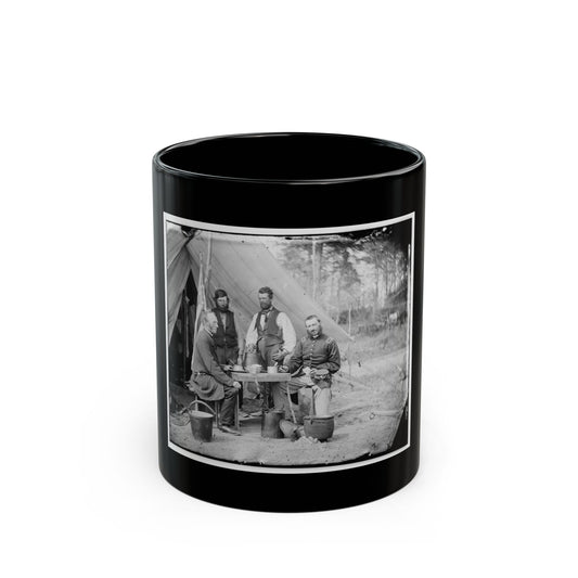 Black-and-white view of four men gathered at a table outside a canvas army tent beside metal kettles.
