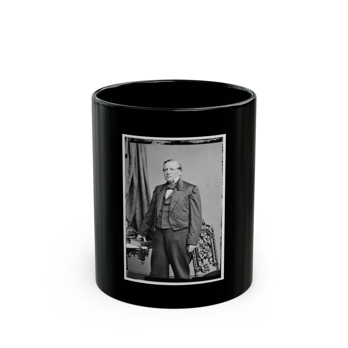 Black and white studio portrait of a standing elderly man in a dark frock coat beside an ornate carved chair.