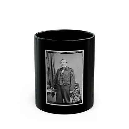 Black and white studio portrait of a standing elderly man in a dark frock coat beside an ornate carved chair.