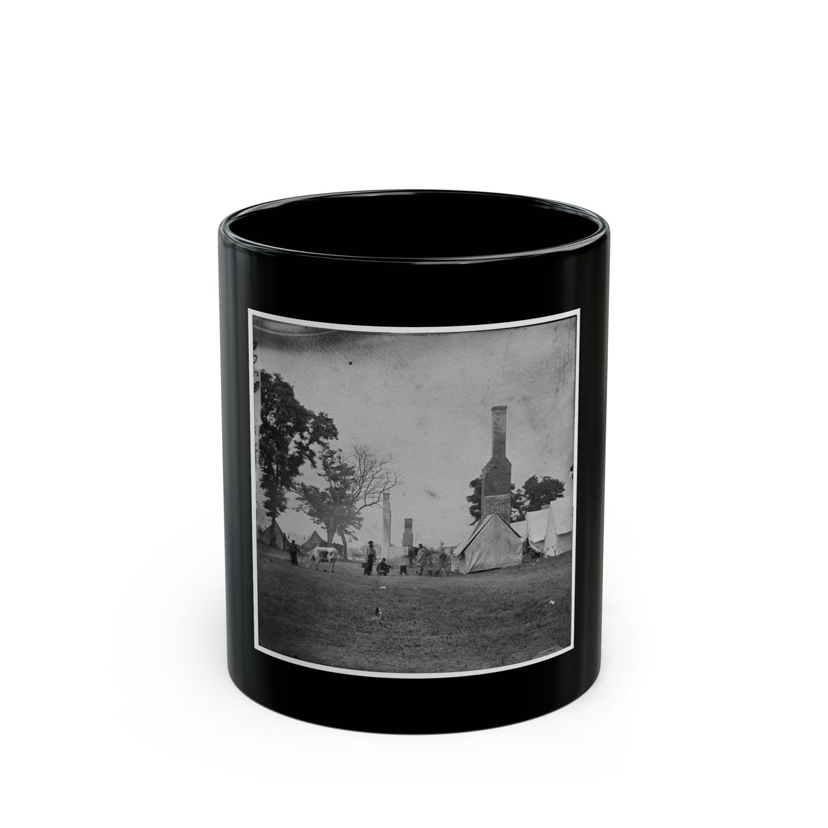 Black and white view of canvas tents and tall brick chimneys standing in an open grassy camp, with a grazing cow.