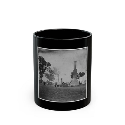 Black and white view of canvas tents and tall brick chimneys standing in an open grassy camp, with a grazing cow.