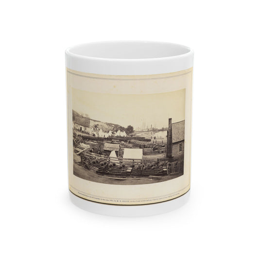 Sepia print of a riverbank camp with shells stacked in racks, wagons, white tents and wooden buildings.