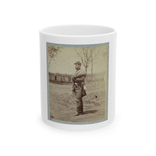 Sepia view of a standing soldier in a kepi holding a sabre, with a picket fence and bare trees behind.