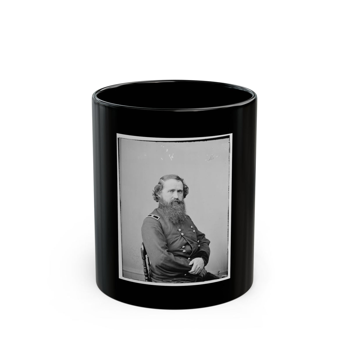 Black and white studio portrait of a bearded man in a dark military coat seated on a wooden chair.