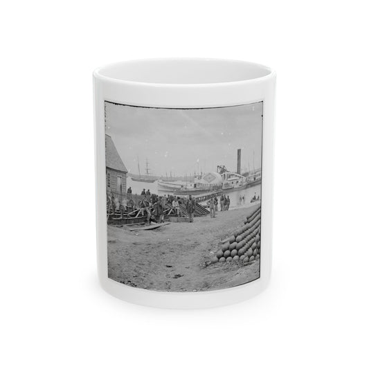 Black-and-white view of soldiers on a harbour dock beside stacked cannonballs, with steamboats on the water.