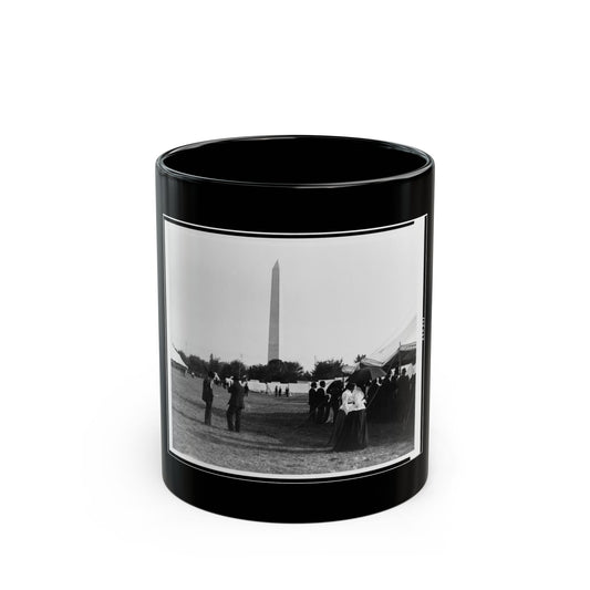Black and white view of a tall obelisk rising above white tents and a crowd of people on a grassy field.