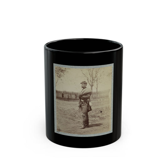 Sepia view of a standing soldier in a kepi holding a sabre, with a picket fence and bare trees behind.