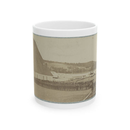 Sepia print showing a long line of soldiers in formation beside a barn-like building, with a steamship on the water.