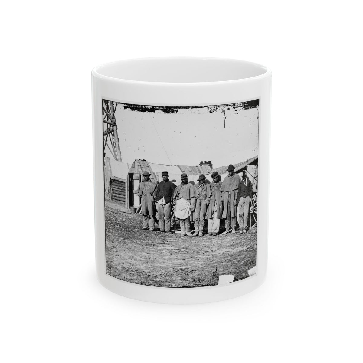 Black and white view of six men standing in a row in front of rustic wooden huts beside a wagon wheel.