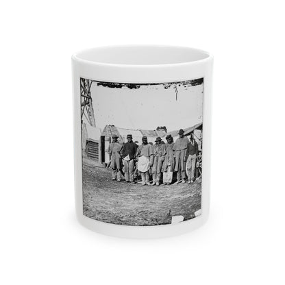 Black and white view of six men standing in a row in front of rustic wooden huts beside a wagon wheel.