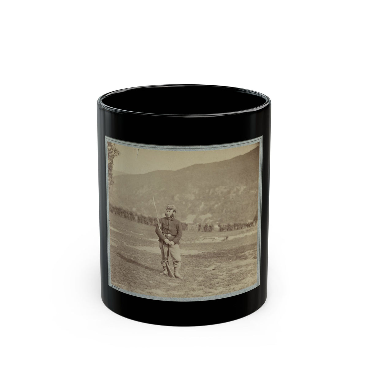 Sepia print of a bearded soldier with a bayoneted rifle standing in a field before a wooded mountain.