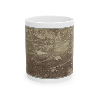 Sepia print of a fallen soldier lying on grass among rocks, with a rifle and a hat on the ground nearby.