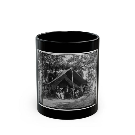 Black and white view of three seated men before a wall tent among tall trees, with laundry hung nearby.