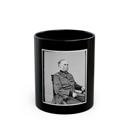 Black and white studio portrait of an older man in a dark button-front coat, seated in a carved wooden chair.