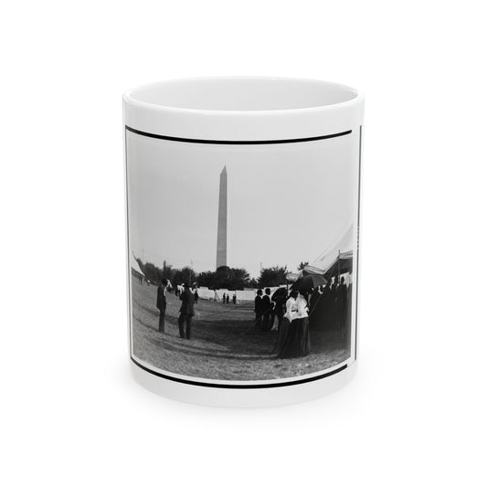 Black and white view of a tall obelisk rising above white tents and a crowd of people on a grassy field.