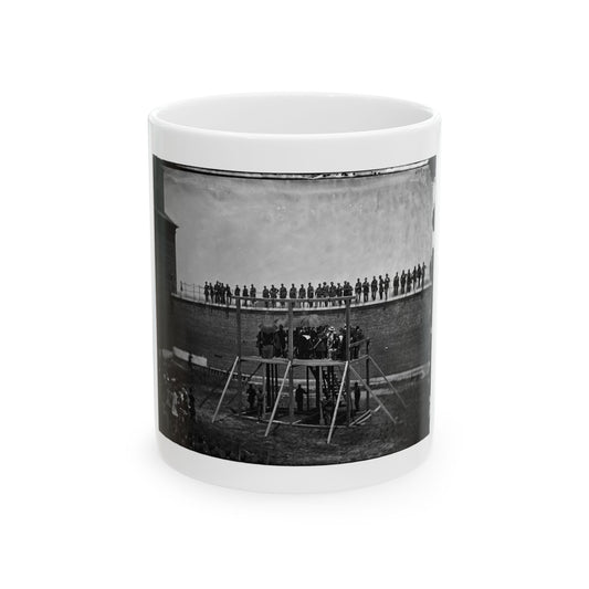 Black and white 1860s view of a timber scaffold before a long brick wall, with soldiers on top and a crowd below.