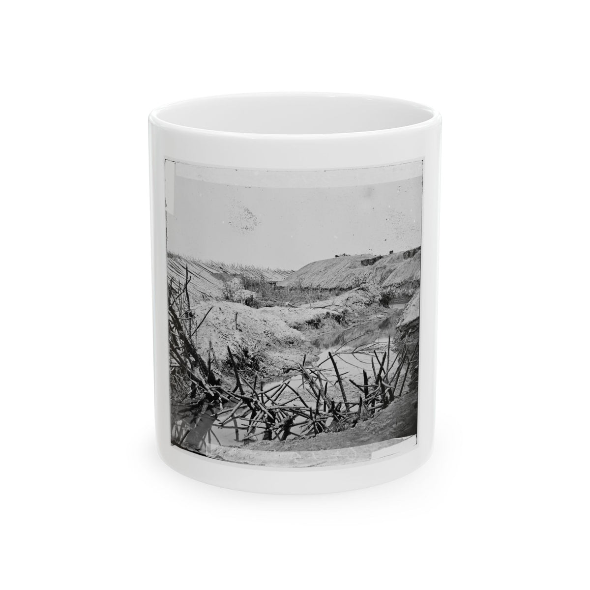 Black and white view of tangled branches in a shallow creek beside dirt embankments and sharpened stakes.