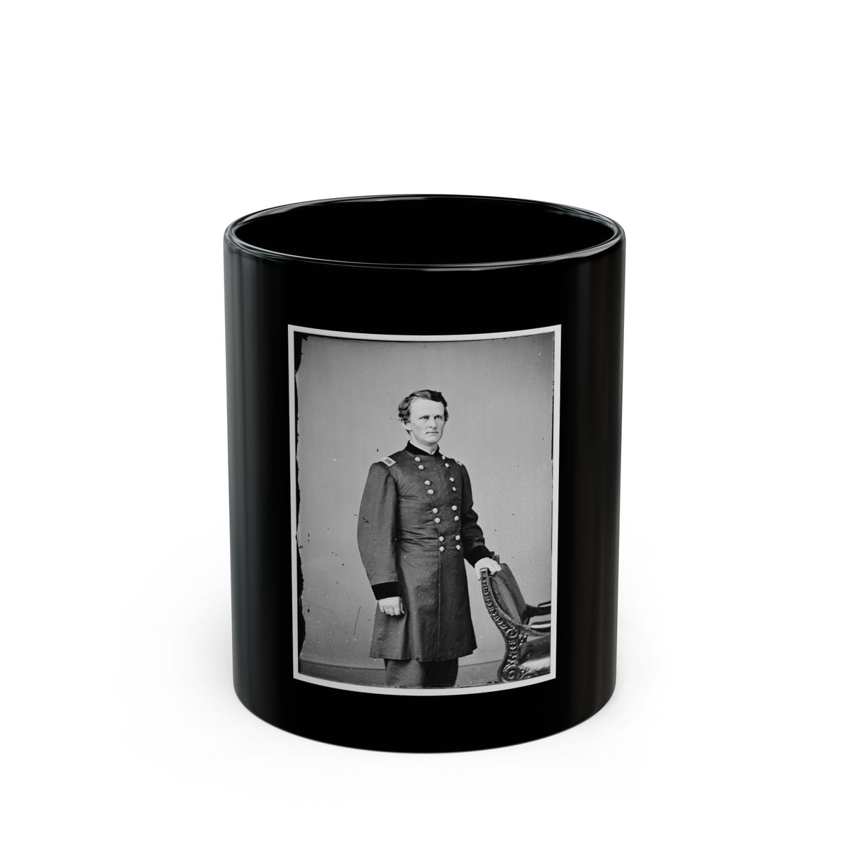 Black and white studio portrait of a young man in a long military coat beside an ornate carved chair.