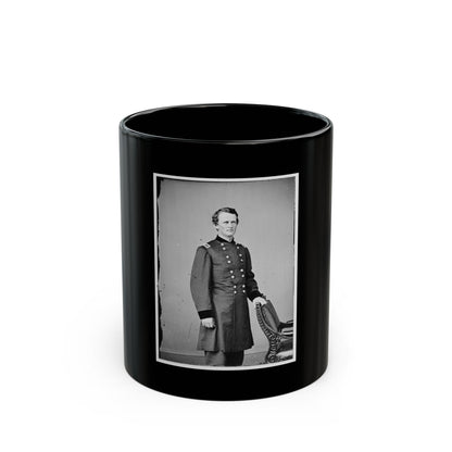 Black and white studio portrait of a young man in a long military coat beside an ornate carved chair.