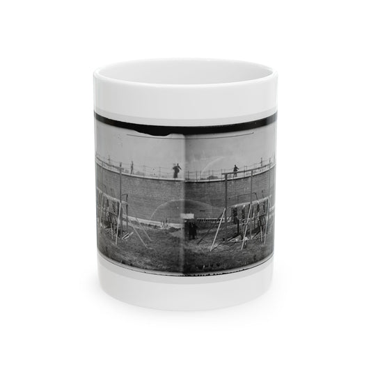 Black and white stereo view of a wooden gallows in a grassy yard beside a tall brick wall, with onlookers above.
