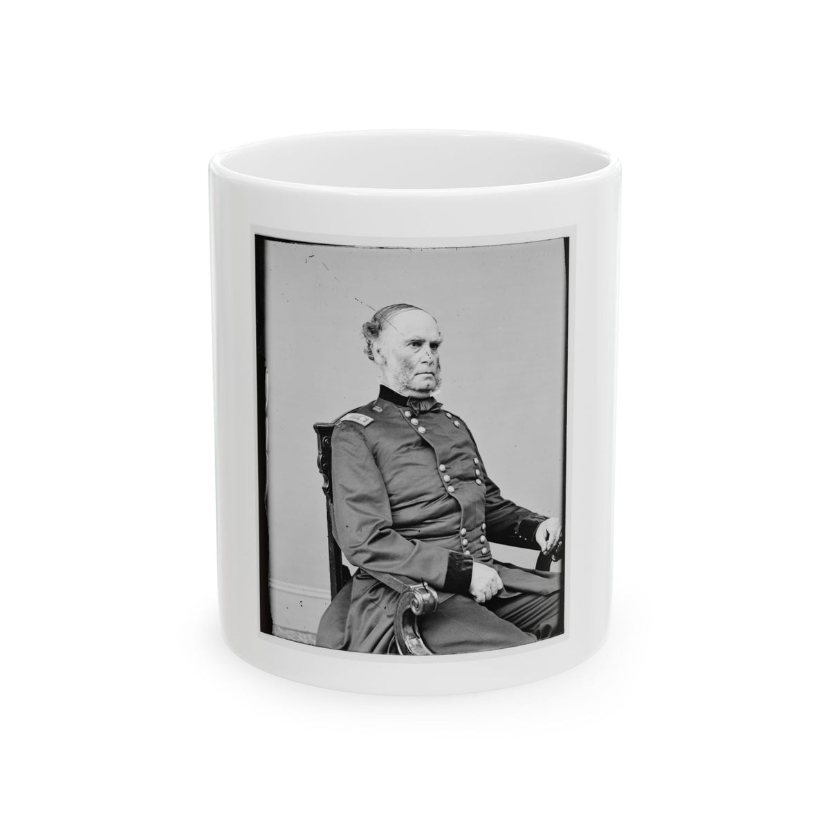 Black and white studio portrait of an older man in a dark button-front coat, seated in a carved wooden chair.