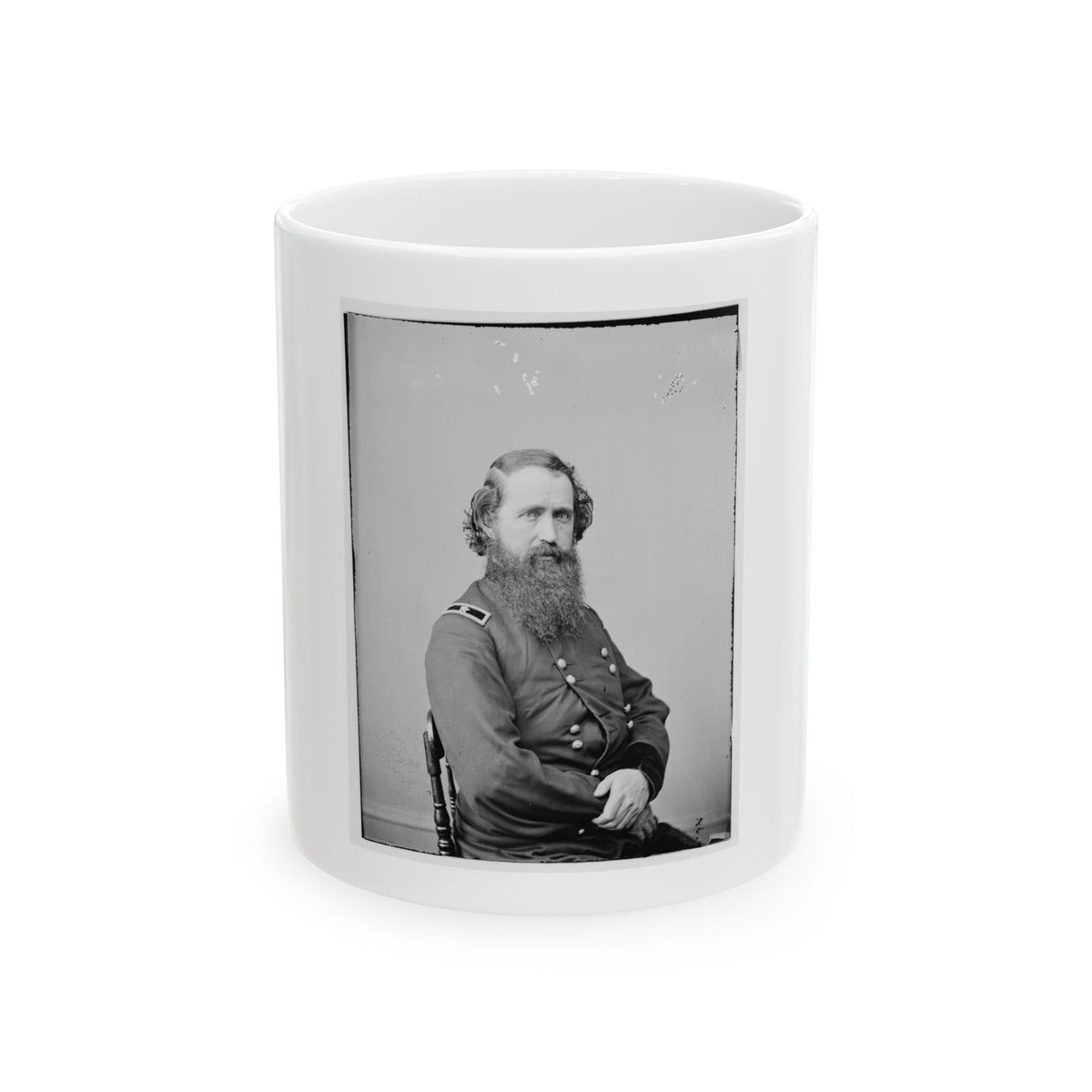 Black and white studio portrait of a bearded man in a dark military coat seated on a wooden chair.