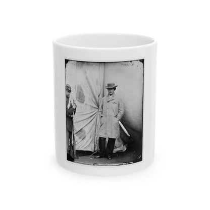 Black and white 1860s studio view of a man in a long coat and hat beside a rifle-holding soldier before draped canvas.