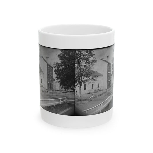 Black-and-white stereograph of whitewashed brick buildings and trees along a dirt street behind a white fence.