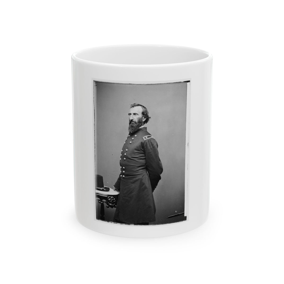 Black and white vintage portrait of a bearded man in a long military coat standing beside a small table with a hat.