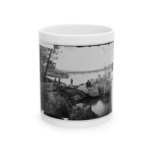 Black and white 19th-century view of men on rocks by a river, with a ferry and stone arch bridge behind.