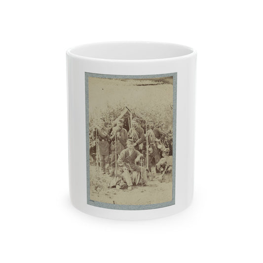 Sepia view of soldiers with rifles and canteens at a leafy lean-to camp, a drummer seated in front.