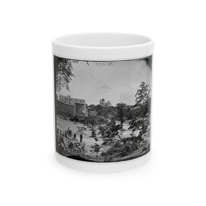 Black-and-white view of a brick mill and waterwheel beside rapids, with men standing on rocks in the stream.