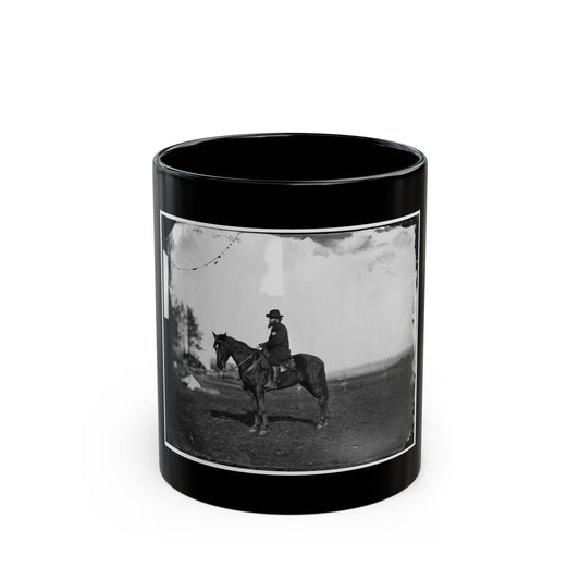 Black and white scene of a bearded rider in a hat and dark coat seated on a dark horse in an open field.