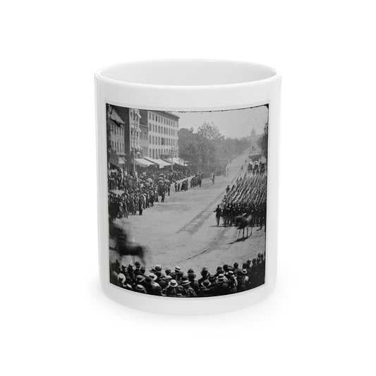 Black and white 19th-century view of marching soldiers with upright rifles on a broad avenue lined with spectators.