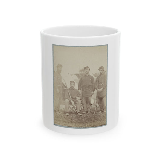 Sepia-toned print of four uniformed soldiers in forage caps posing with rifles on a grassy camp, one seated.