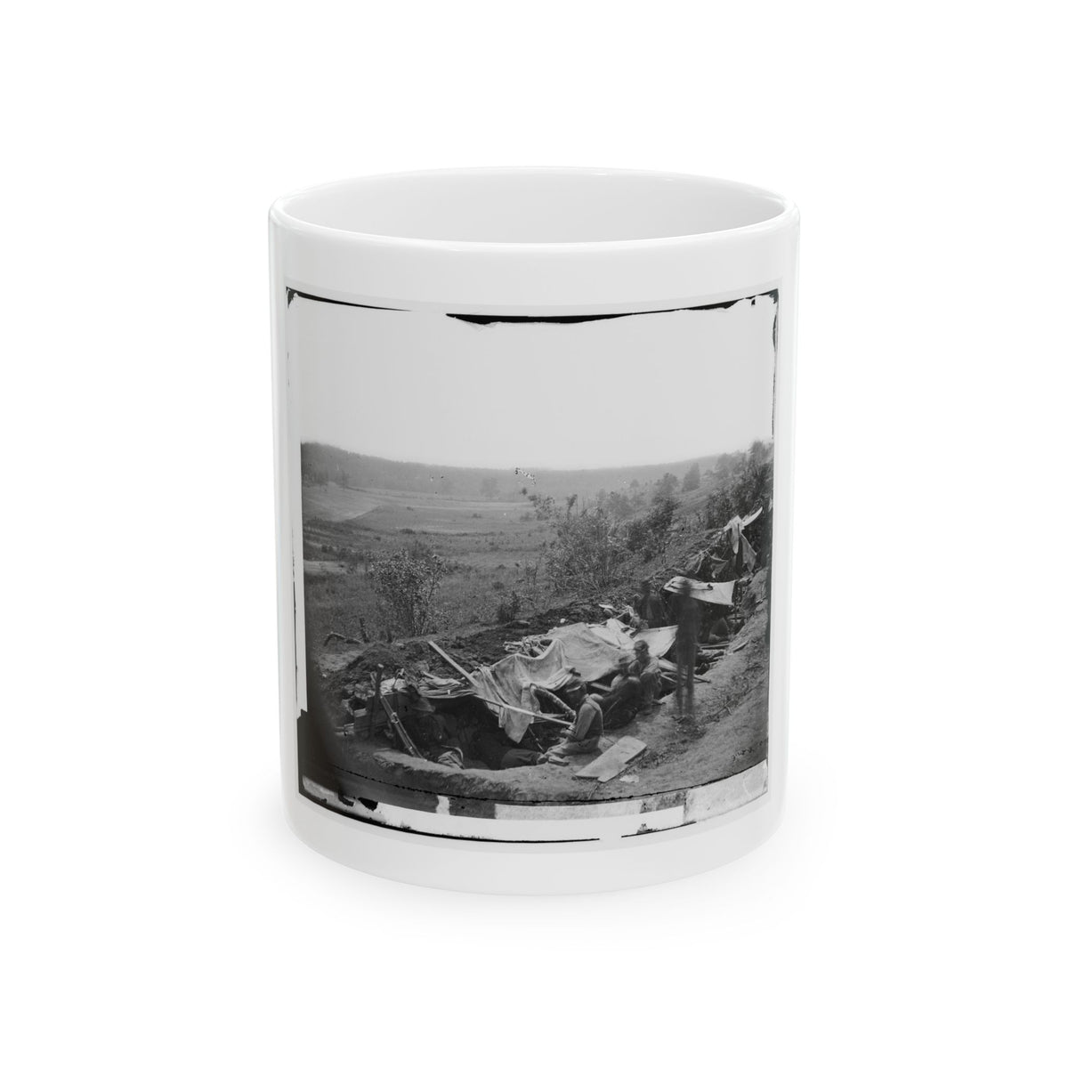 Black and white view of soldiers sheltering under canvas in a dug-in earthwork overlooking an open field.