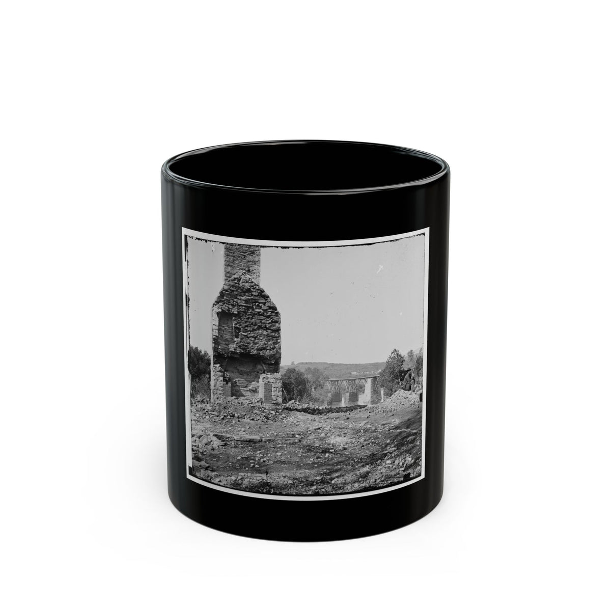 Black and white view of a ruined stone chimney rising above rubble, with a wooden trestle bridge beyond.