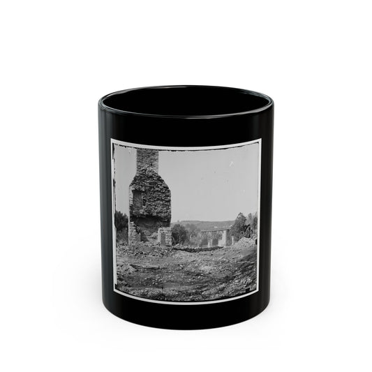Black and white view of a ruined stone chimney rising above rubble, with a wooden trestle bridge beyond.