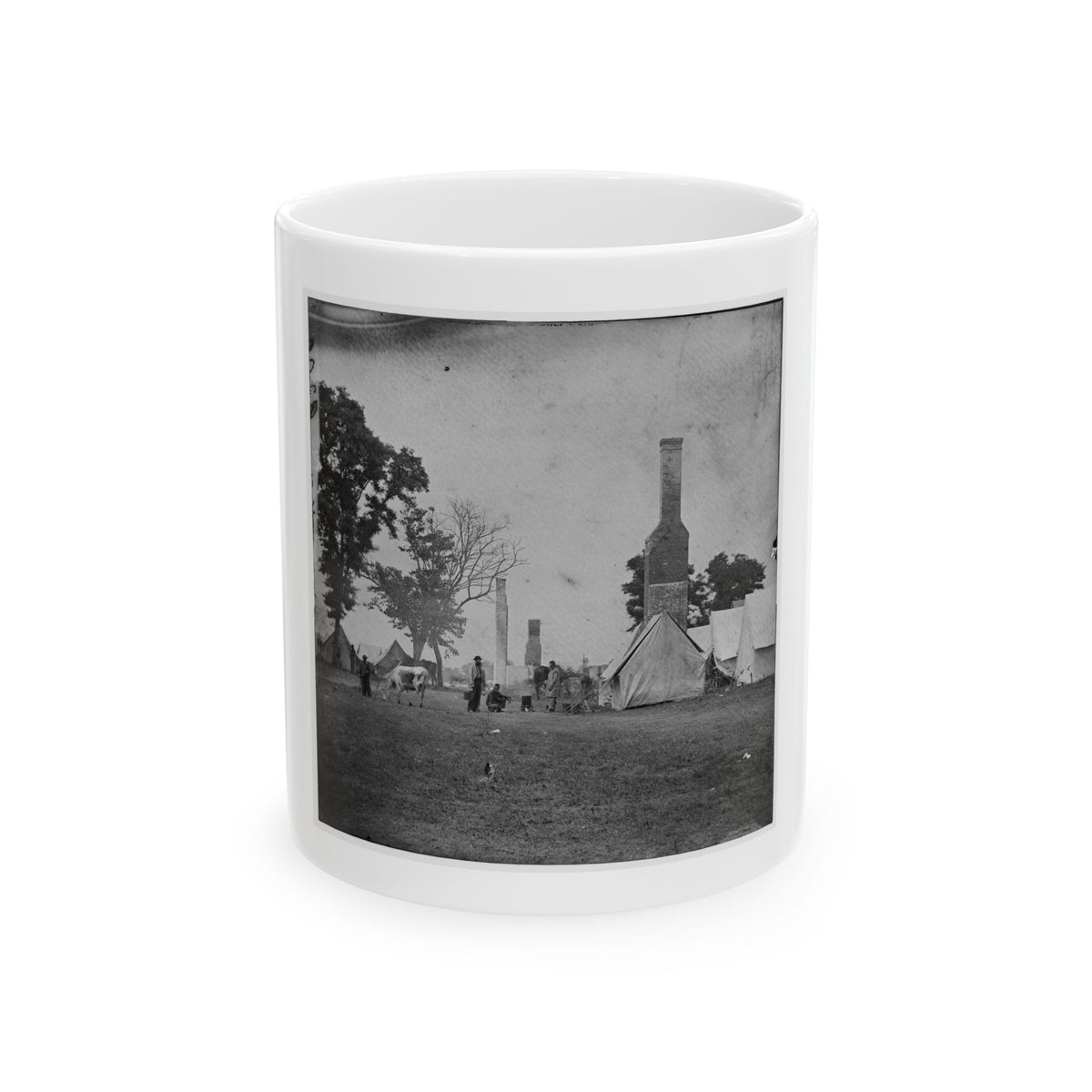 Black and white view of canvas tents and tall brick chimneys standing in an open grassy camp, with a grazing cow.