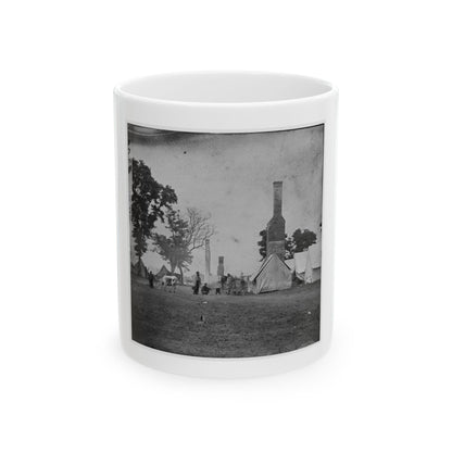 Black and white view of canvas tents and tall brick chimneys standing in an open grassy camp, with a grazing cow.