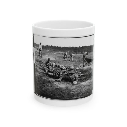 Black and white 1860s print of skulls and remains on a wooden litter in a grassy field, with men digging nearby.