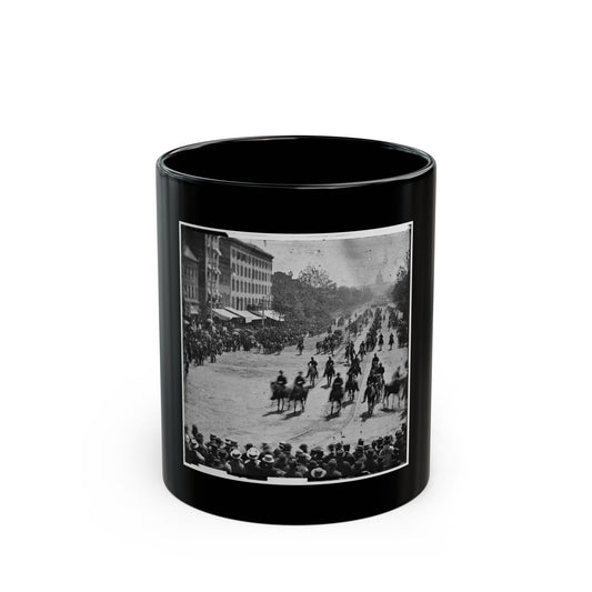 Black and white historical view of mounted cavalry riding down a tree-lined city avenue past a crowd of onlookers.