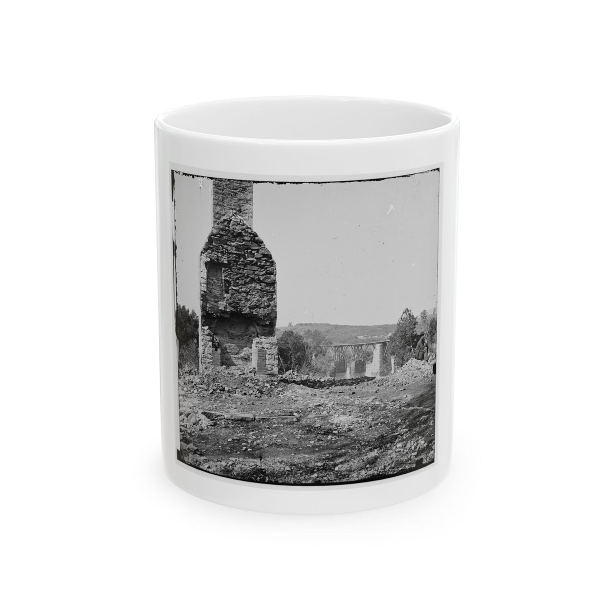 Black and white view of a ruined stone chimney rising above rubble, with a wooden trestle bridge beyond.