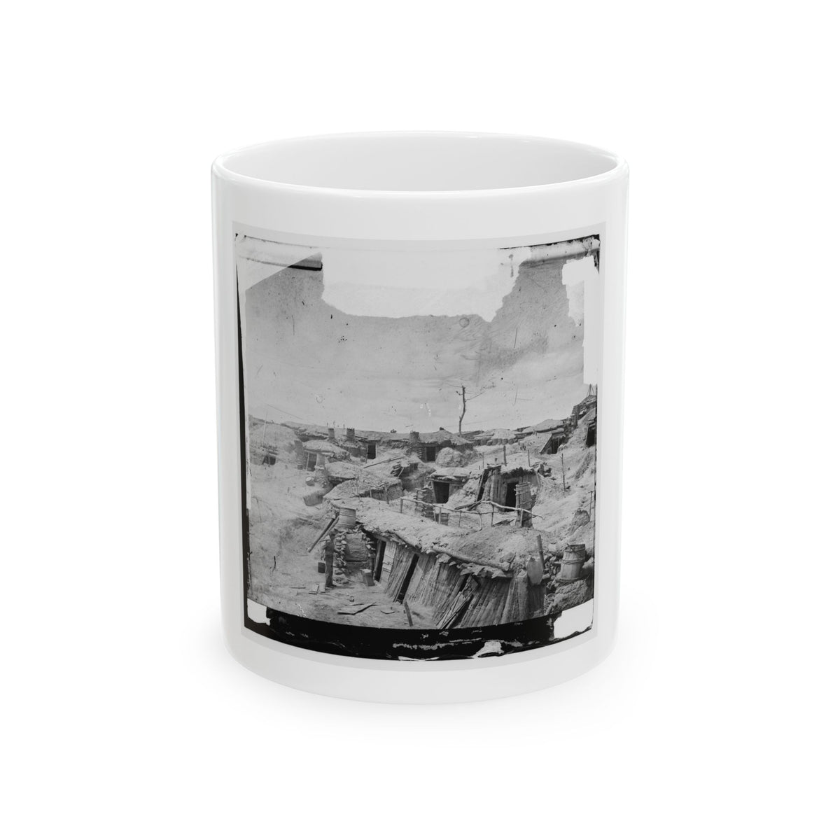 Black-and-white view of a dugout camp of earth-roofed shelters with wooden doors, barrels and one standing figure.
