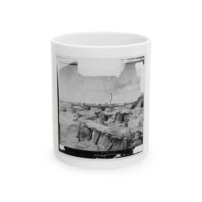 Black-and-white view of a dugout camp of earth-roofed shelters with wooden doors, barrels and one standing figure.