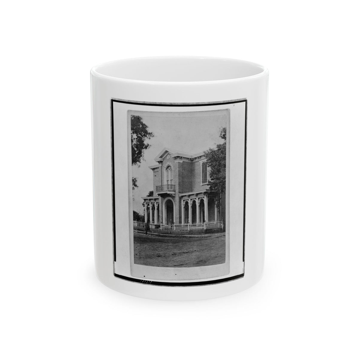 Black and white view of a brick Italianate house with arched windows, a columned porch and picket fence.